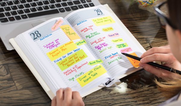 Close-up Of A Businessperson Writing Schedule In Diary On Wooden Desk